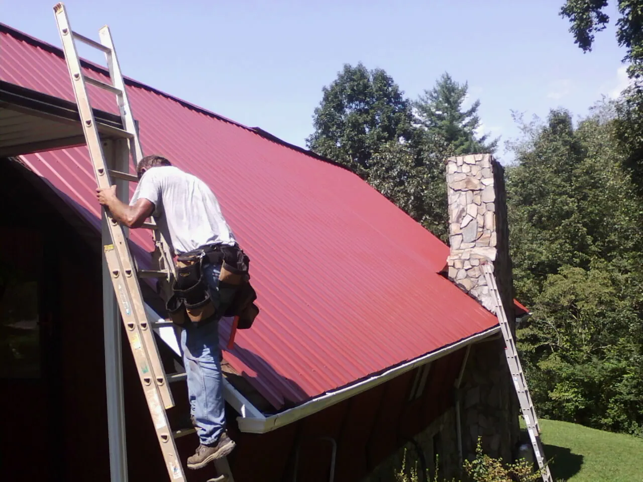 Roofer on a ladder beside a red metal roof and stone chimney — Soffit & Fascia Repair by Roofing Brothers in Morgantown